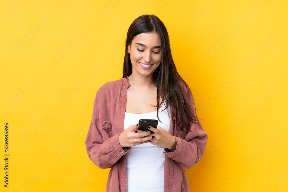 Young brunette woman over isolated yellow background sending a message with the mobile Young brunette woman over isolated yellow background sending a message with the mobile