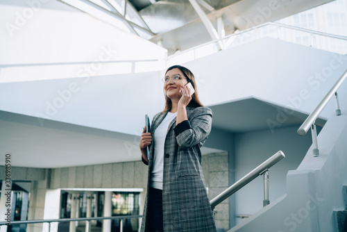 Smiling hipster girl in trendy outfit and eyewear making mobile phone call standing on urban architecture stairs, positive caucasian woman satisfied with roaming connection talking on smartphone