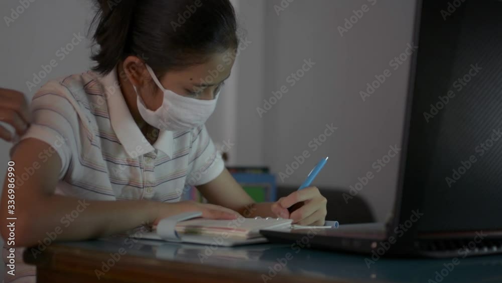 Asian cute girl wearing protective mask studying from home with video call during COVID-19 pandemic situation. Mother wearing protective mask approaching and teaching her daughter study from home. Asian cute girl wearing protective mask studying from home with video call during COVID-19 pandemic situation. Mother wearing protective mask approaching and teaching her daughter study from home.