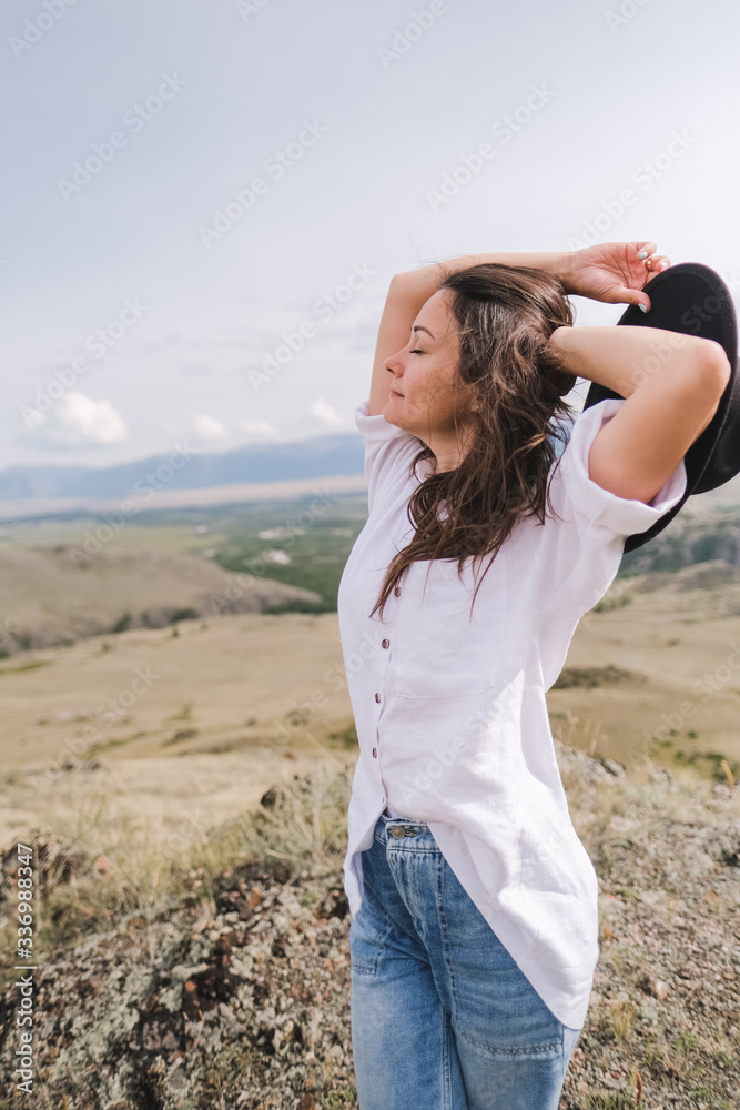 Attractive brunette in a hat and white shirt is enjoying nature in the mountains. Beautiful young woman in a shirt and jeans walks along the beautiful landscape Attractive brunette in a hat and white shirt is enjoying nature in the mountains. Beautiful young woman in a shirt and jeans walks along the beautiful landscape