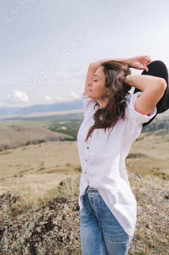 Attractive brunette in a hat and white shirt is enjoying nature in the mountains. Beautiful young woman in a shirt and jeans walks along the beautiful landscape