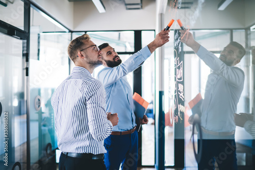 Confident male employee pointing on notes standing with colleague near glass creating infographic for conversation, skilled managers discussing sketches and planning during informal meeting in office photo