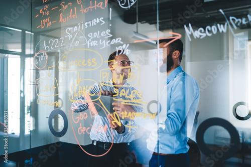 Cheerful partners smiling while collaborating near modern glass board with statistics planning for business marketing, entrepreneurs creating strategy indoors discussing web publication on gadget photo