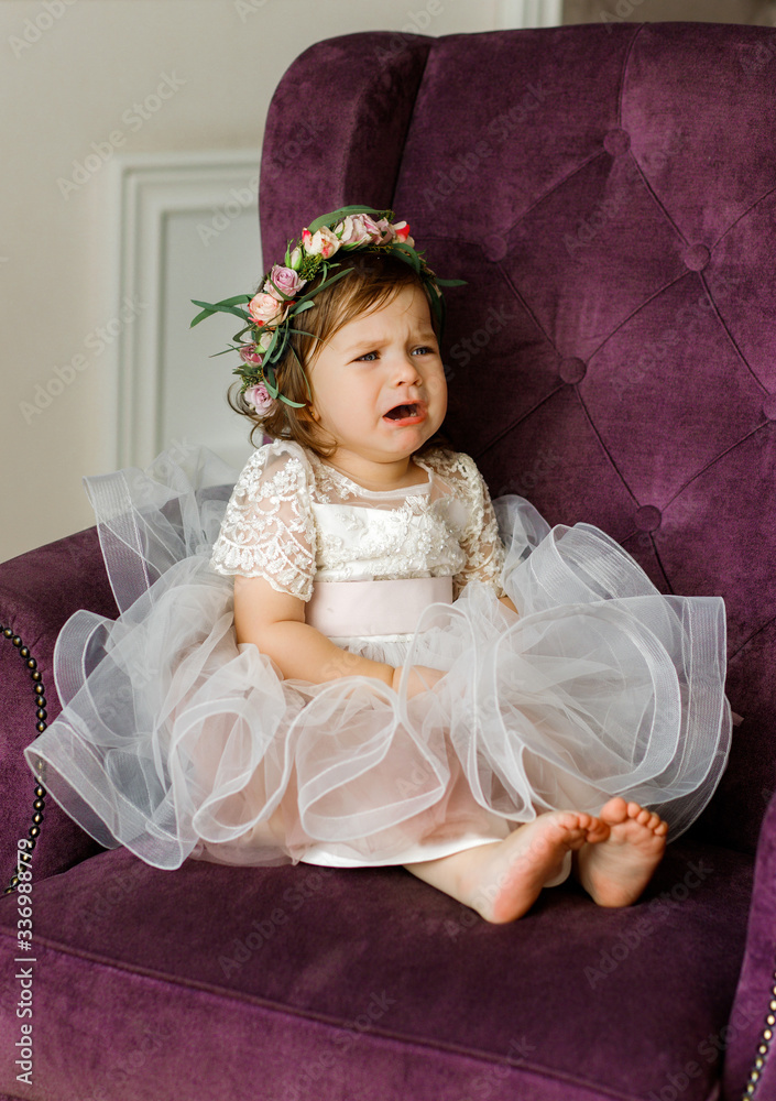 Fototapeta premium Emotional little girl in a pink dress and floral wreath sits on a chair.