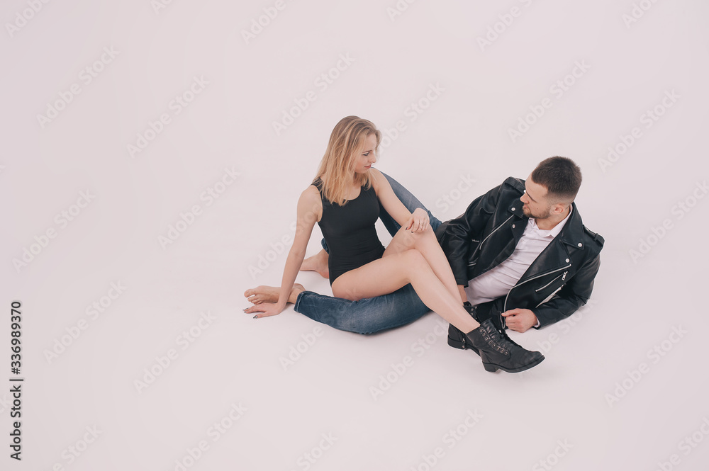 Happy time. Beautiful young couple sitting on floor in studio. Man wearing in denim clothes and leather jacket. Beautiful young sexy girl in black bodysuit and shoes. Happy time. Beautiful young couple sitting on floor in studio. Man wearing in denim clothes and leather jacket. Beautiful young sexy girl in black bodysuit and shoes.