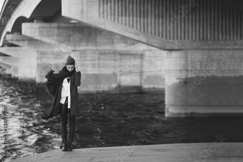 Beautiful lonely woman in a coat and scarf developing in the wind under a bridge near the river. The concept of loneliness. Black and white art photo. Soft focus.