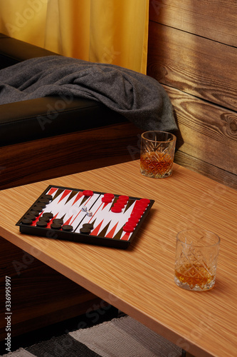 Staged photo of backgammon on the bedside table with whiskey glasses near the couch and a yellow curtain. The narde set consists of a folding board, black and red playing chips and two cubes.  photo