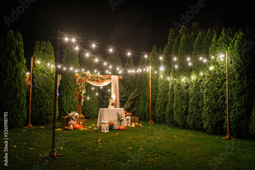 night wedding ceremony, the arch is decorated with flowers, candles and garlands of light bulbs and there is a wedding cake on the table