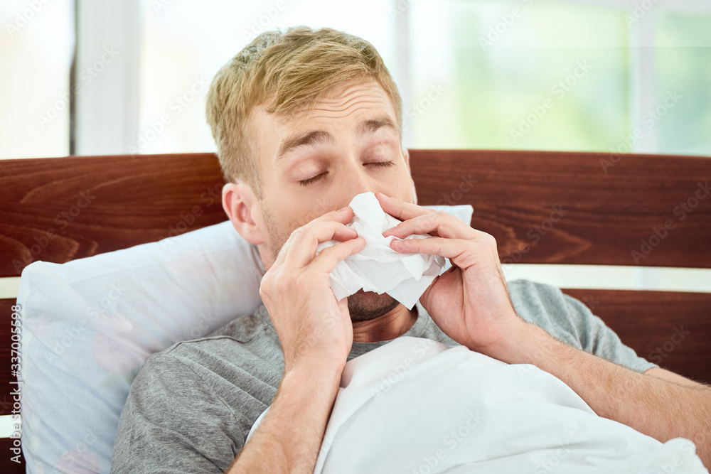 Portrait of a sick young man with runny nose suffering from cold or flu and sneezing while lying in bed at home. Virus disease Portrait of a sick young man with runny nose suffering from cold or flu and sneezing while lying in bed at home. Virus disease