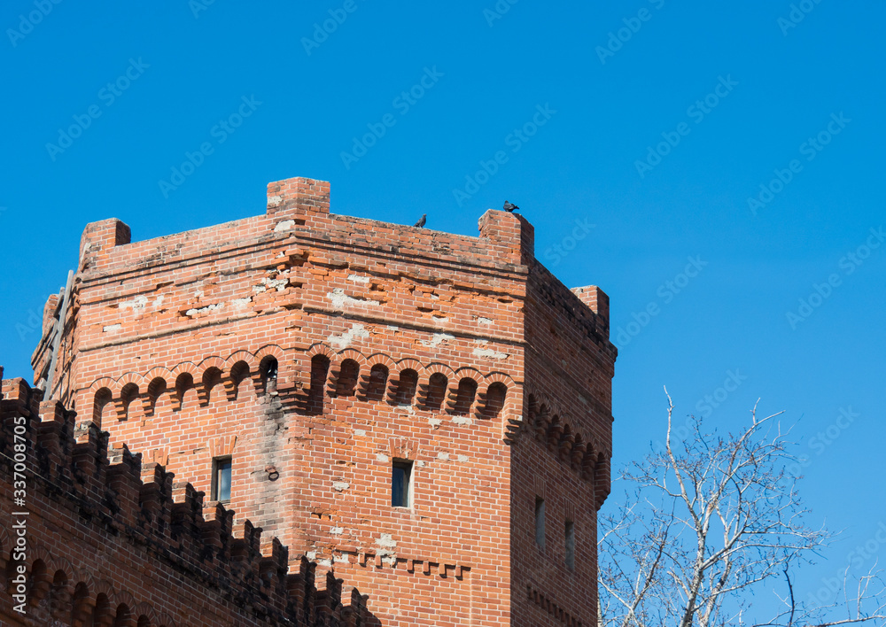 Red brick fortress tower with loopholes and a piece of fence against a blue sky.