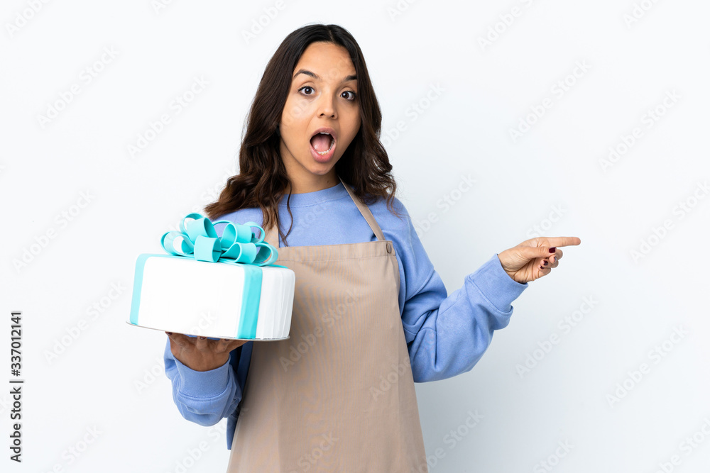 Pastry chef holding a big cake over isolated white background surprised and pointing side Pastry chef holding a big cake over isolated white background surprised and pointing side