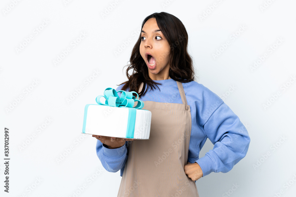 Pastry chef holding a big cake over isolated white background doing surprise gesture while looking to the side Pastry chef holding a big cake over isolated white background doing surprise gesture while looking to the side