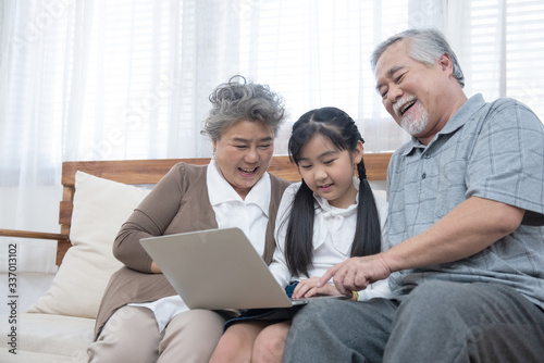 Happy asian grandparent with little young cute grandchild sitting on sofa playing laptop together.