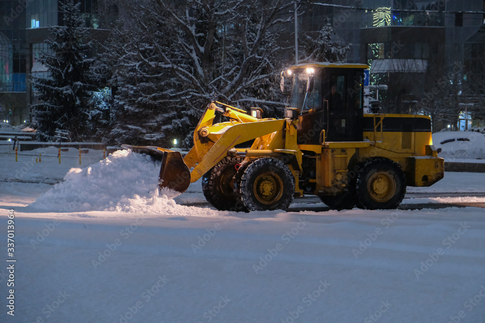 Wheel loader is removing snow at night during snowfall. Selective focus, blurred. Snow removing concept. Wheel loader is removing snow at night during snowfall. Selective focus, blurred. Snow removing concept.