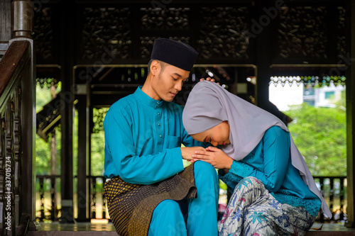 Young couple of malay muslim in traditional costume showing apologize gesture during Aidilfitri celebration at traditional wooden house.