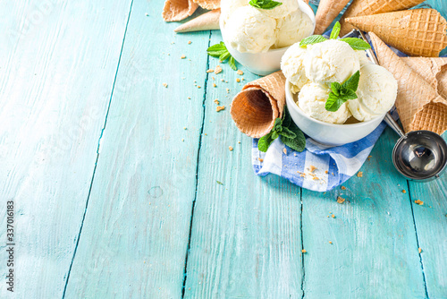 Homemade white vanilla ice cream balls with mint leaves in small portioned white bowl. With ice cream waffle cone and spoon,  on rustic turquoise wooden table, copy space