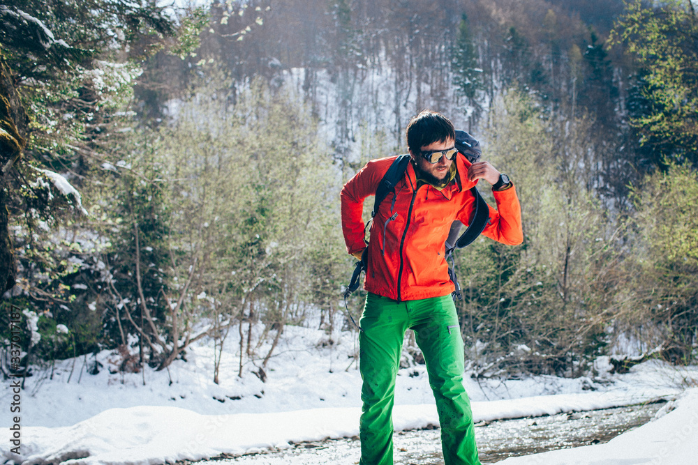 Young male tourist takes off his backpack near a river in the mountains..Beautiful winter landscape with snow covered banks and trees  on background. Climbing, hiking, trekking, active life concept.