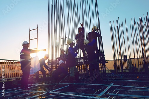 The construction of a rooftop high rise building, workers are tied with steel bars for concrete pouring, surveying the site and cranes are lifting or moving metal at the construction site. Pastel tone