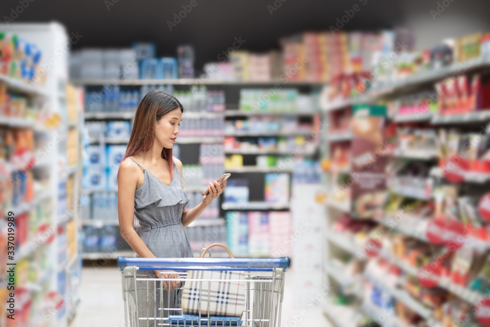 image of asian shopper woman leaning on shopping cart, using a mobile phone and smiling, in the background choosing food