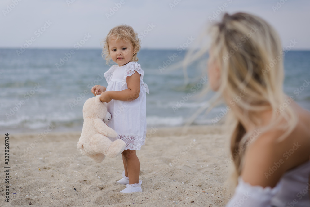 A blonde girl holds a toy bear and looks at her mother by the blue sea at sunset. A blonde girl holds a toy bear and looks at her mother by the blue sea at sunset.