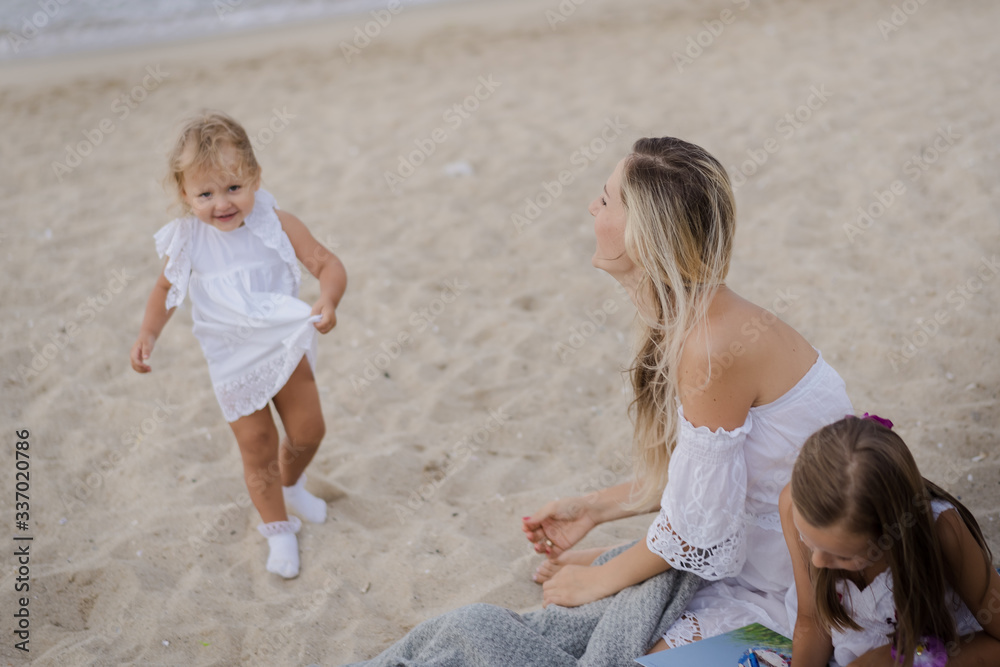 Mom with daughters blondes in white dresses laugh, hug and sit near the blue sea on the beach at sunset. Mom with daughters blondes in white dresses laugh, hug and sit near the blue sea on the beach at sunset.