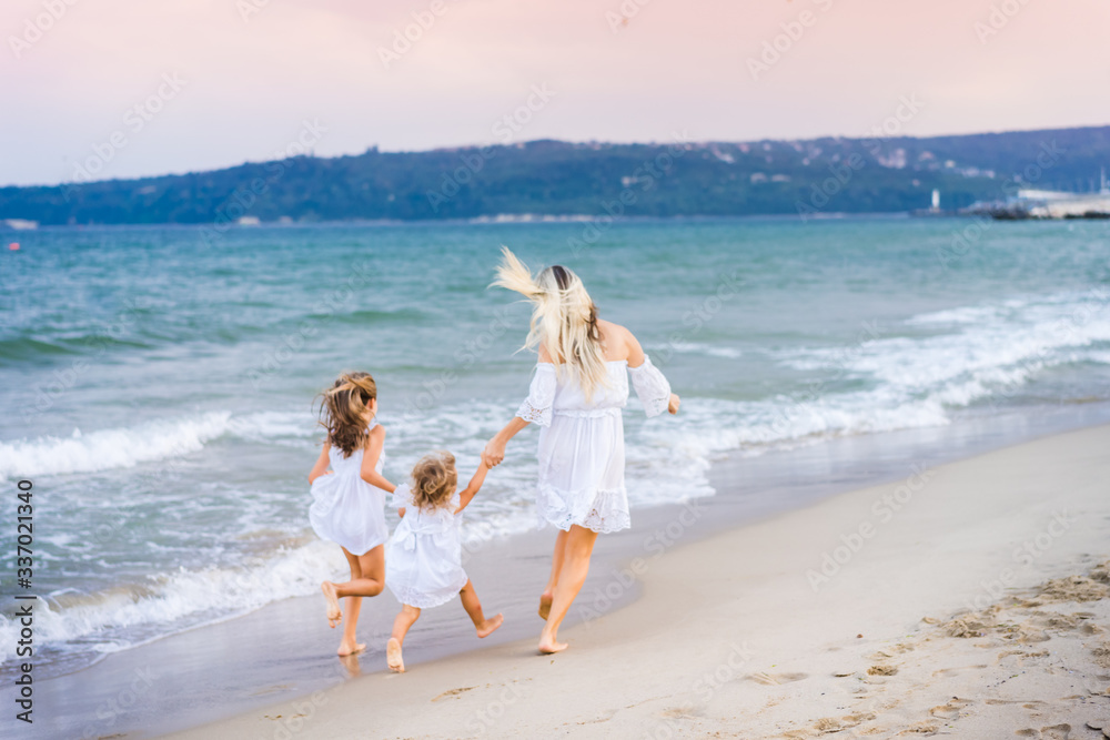 Blonde mother and two daughters in white dresses run near the blue sea and laugh on the beach at sunset. Back view. Blurred. Blonde mother and two daughters in white dresses run near the blue sea and laugh on the beach at sunset. Back view. Blurred.