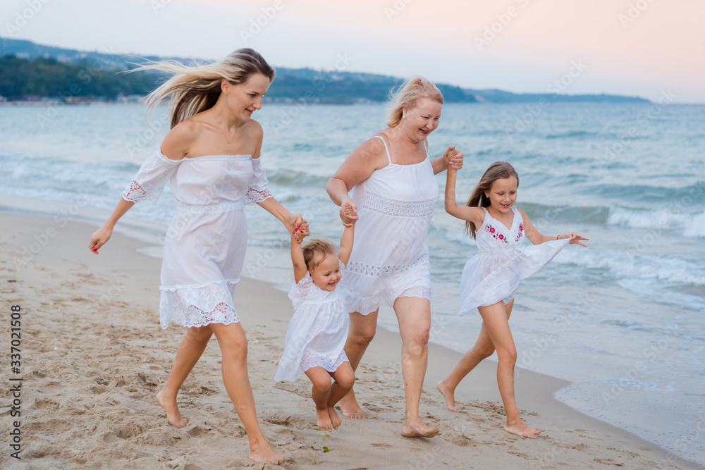 Blondes grandmother, mother and two granddaughters in white dresses run near the blue ocean and laugh on the beach at sunset.