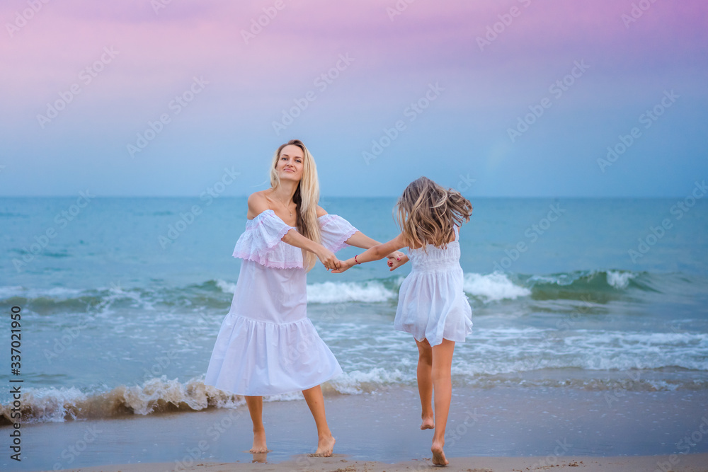 Mom and daughter blondes in white dresses laugh, hug and roundelay near the blue sea on the beach at sunset.