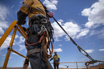 Safe workplace rigger wearing working at heights harness clipping an inertia reel shock absorbing fall arrest device hook on the rope sling anchor point while working from 2 metre exposure open edges 
