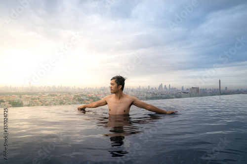 Asian young man relaxing in the sky swimming pool watching cityscape 