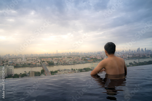Asian young man relaxing in the sky swimming pool watching cityscape 