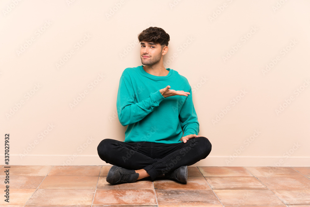 Young handsome man sitting on the floor extending hands to the side for inviting to come Young handsome man sitting on the floor extending hands to the side for inviting to come