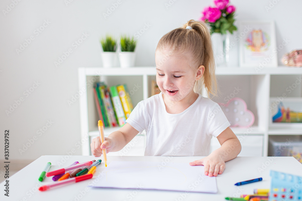 Blonde happy little girl with ponytale drawing and writing lsitting by the white table in light roon. Preschool education, early learning Blonde happy little girl with ponytale drawing and writing lsitting by the white table in light roon. Preschool education, early learning