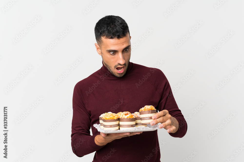 Young man over isolated white background holding mini cakes and surprised Young man over isolated white background holding mini cakes and surprised