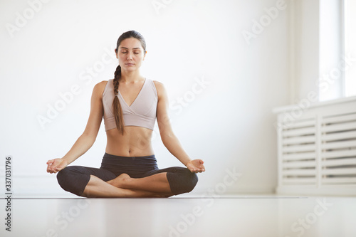 Portrait of young healthy woman sitting in lotus position and meditating in white room