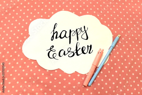 Photo of white paper in the shape of cloud on pink polka dot fabric. Happy Easter - lettering. Color abstract mockup background template with pen pencil marker bunny