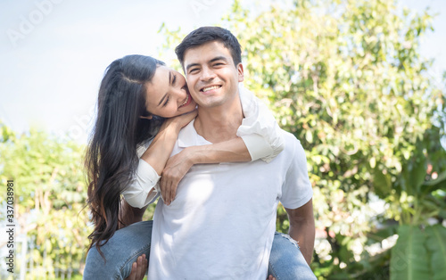 Portrait of cute romantic caucasian guy in checkered shirt, dreamy asian lady rides him on rear. Leisure, chill happiness, lawn stroll, relax, romance lifestyle, well dressed partners posing