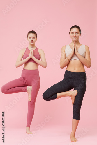 Portrait of two healthy women standing on one leg and doing yoga over pink background