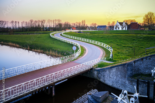 Curvy Road and white fence in old dutch countryside near an old water lock leading towards historic farmhouse © fotografiecor