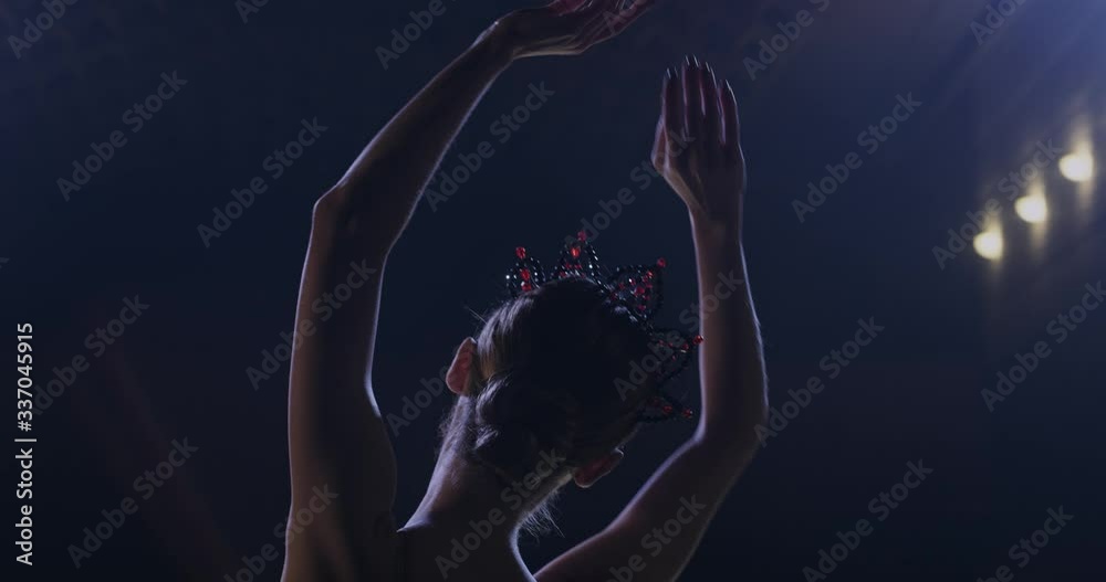 Back view of young pretty Caucasian ballerina in crown rehearsing hands pas in darkness and ligh of lamps. Close up of female ballet dancer dancing gracefully and looking at camera. Rear. Back view of young pretty Caucasian ballerina in crown rehearsing hands pas in darkness and ligh of lamps. Close up of female ballet dancer dancing gracefully and looking at camera. Rear.