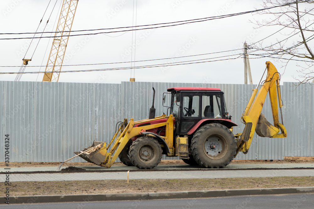 a tractor with two excavator buckets in front and behind on the sidewalk. excavator at a construction site on a background of a metal fence.