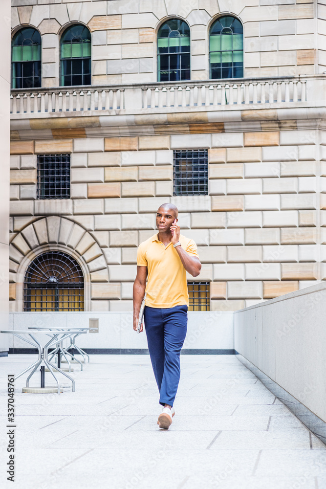 Young African American Man traveling, working in New York City, wearing yellow short sleeve shirt, blue pants, white sneakers, carrying laptop computer, walking outside office, talking on cell phone..