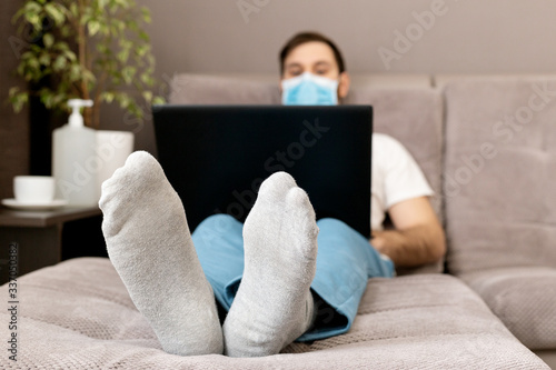 Happy relaxed young caucasian man working from home wearing protective mask using laptop. Cozy Home office, workplace on sofa during coronavirus pandemic, covid 19 quarantine. Remote work, freelancer photo