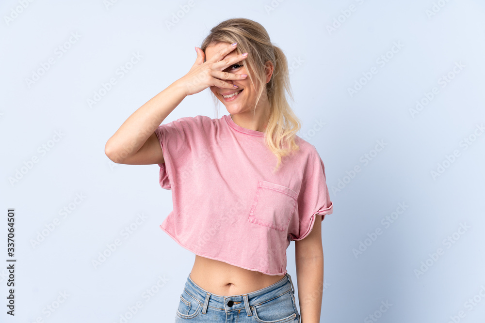 Blonde woman over isolated blue background covering eyes by hands and smiling Blonde woman over isolated blue background covering eyes by hands and smiling