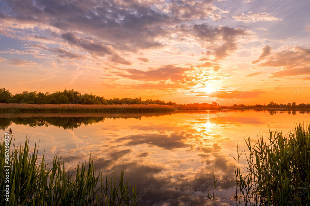 Scenic view of beautiful sunset or sunrise above the pond or lake at spring  or early summer evening with cloudy sky background and reed grass at  foreground. Landscape. Water reflection. Photos
