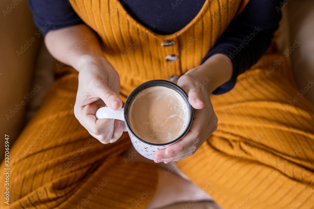 Top view of unrecognizable woman holding a hot cup of coffee at home. Detail of warm cup of latte on white mug. Healthy lifestyle. Woman drinking exotic coffee for balance diet. Beverage concept.