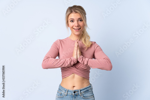 Blonde woman over isolated blue background keeps palm together. Person asks for something