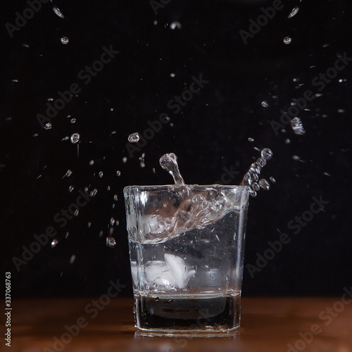 A glass of water on a black background, into which pieces of ice are thrown and splashes fly