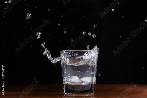 A glass of water on a black background, into which pieces of ice are thrown and splashes fly