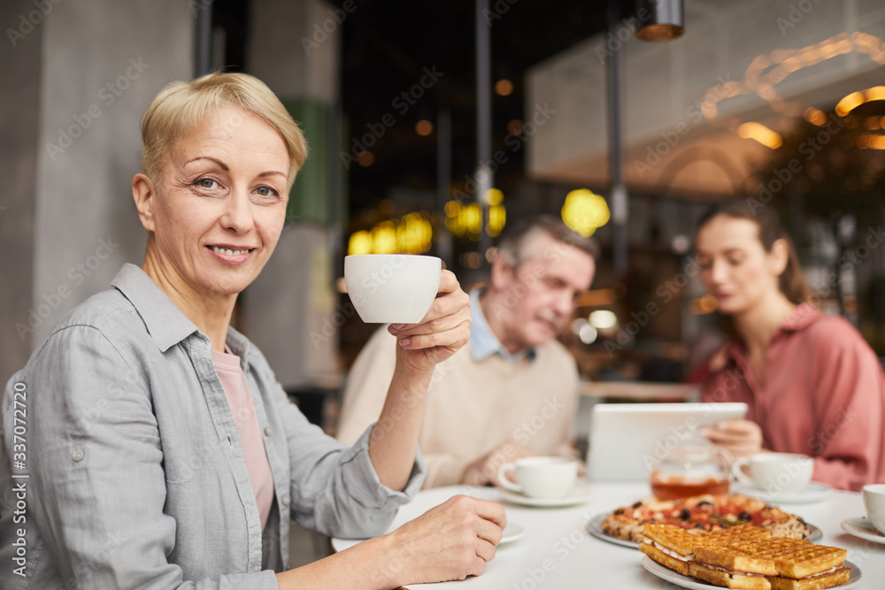 Portrait of mature woman holding cup of tea and smiling at camera while having lunch with her friends in cafe Portrait of mature woman holding cup of tea and smiling at camera while having lunch with her friends in cafe